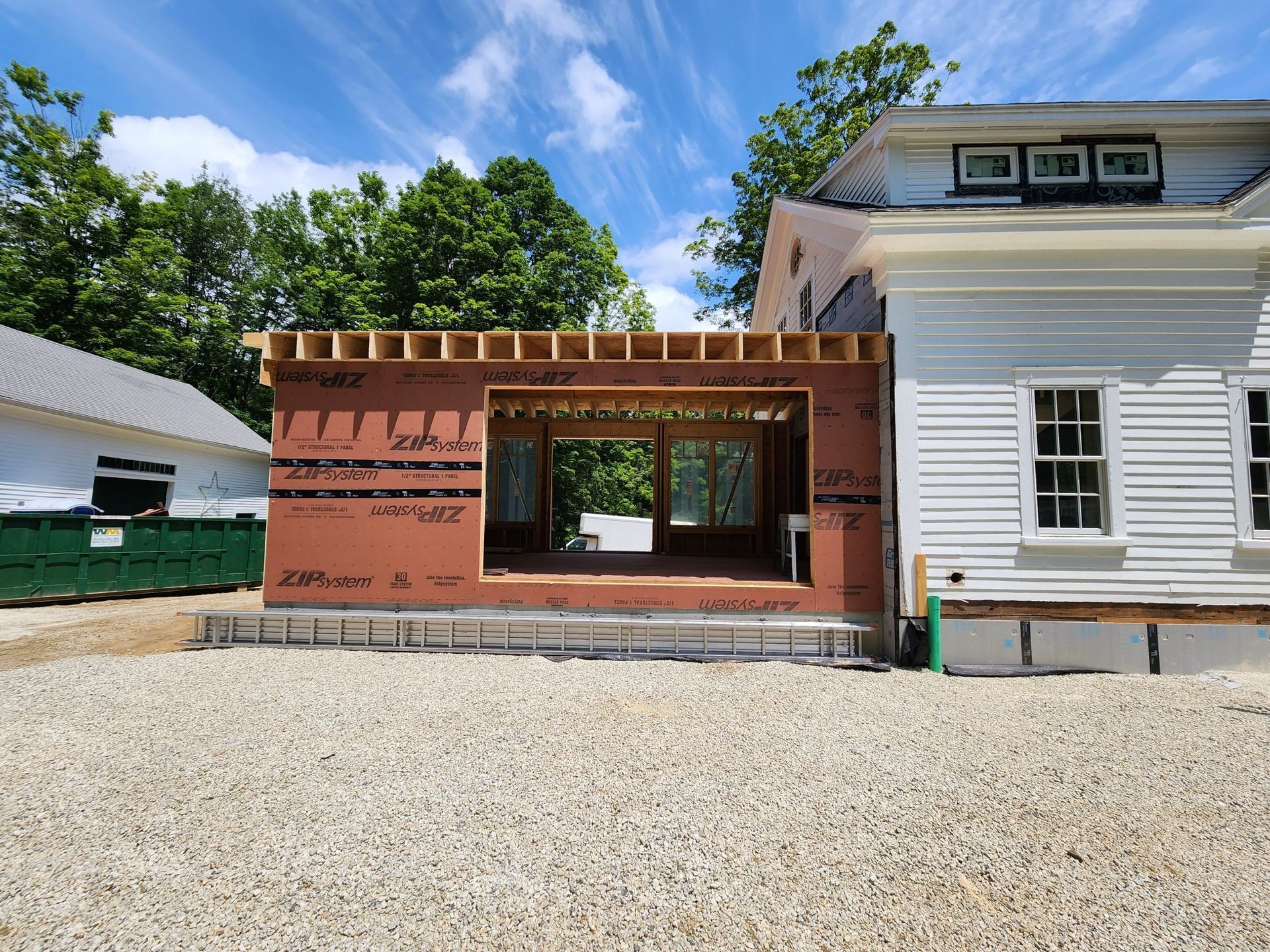 Construction of a new building addition attached to a white house, on a gravel lot.