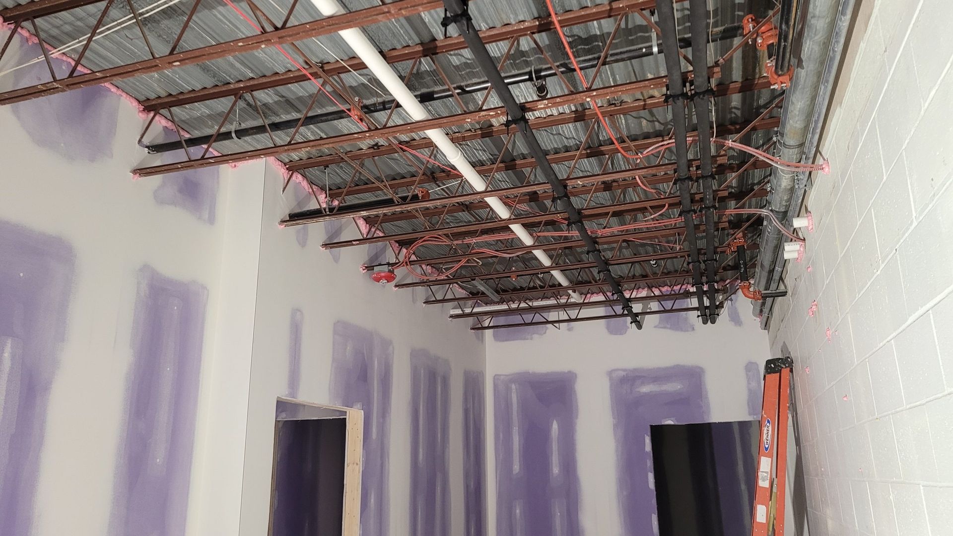 Interior hallway under construction, exposed metal ceiling beams, drywall walls, and a ladder.