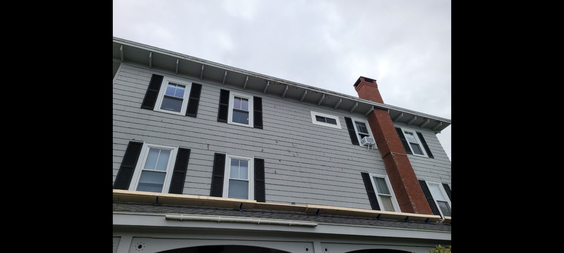 Gray house with black shutters, white trim, and a brick chimney under a cloudy sky.