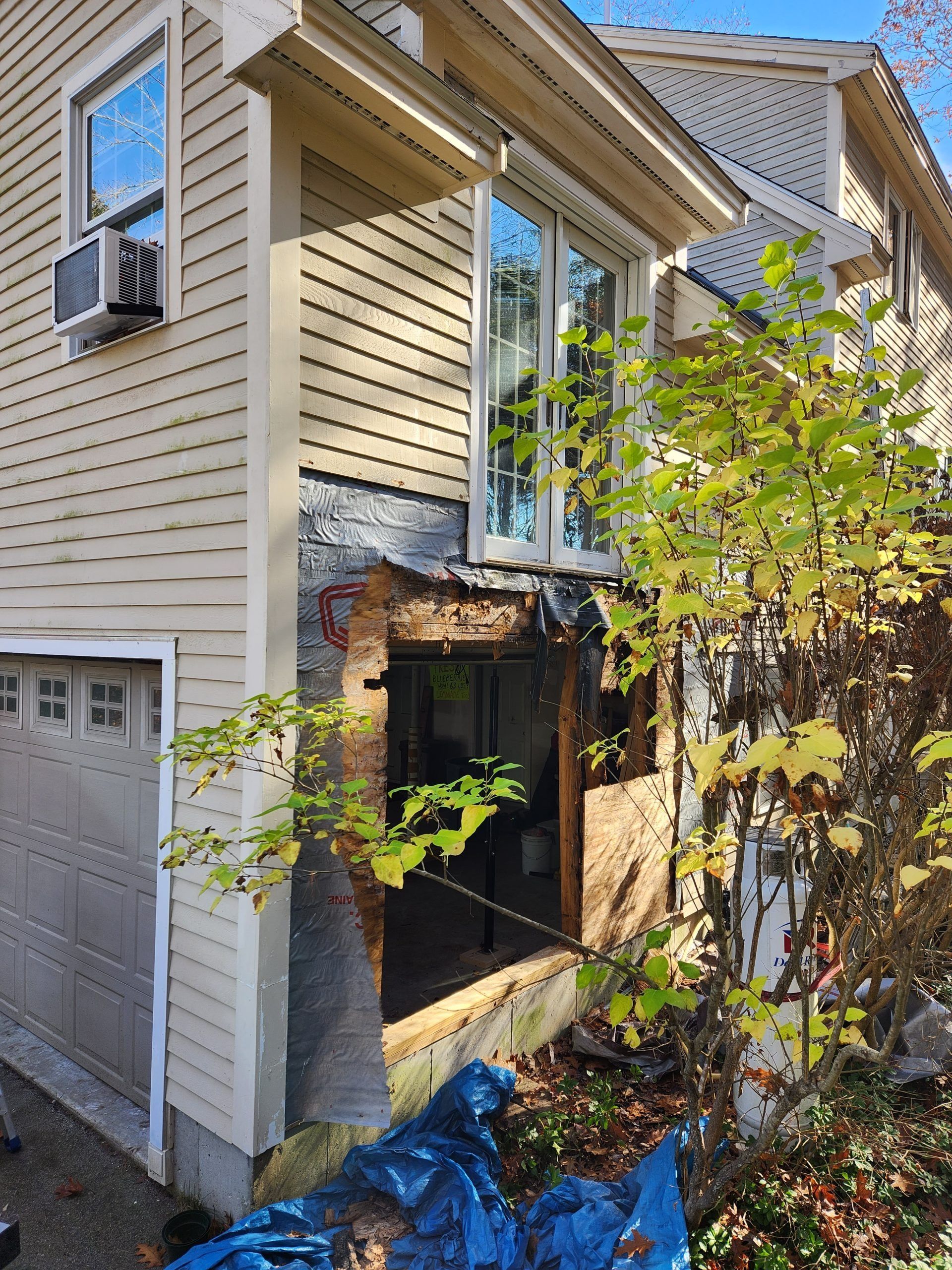 Exterior wall of a house with visible damage and a missing section; beige siding, window, and garage door.