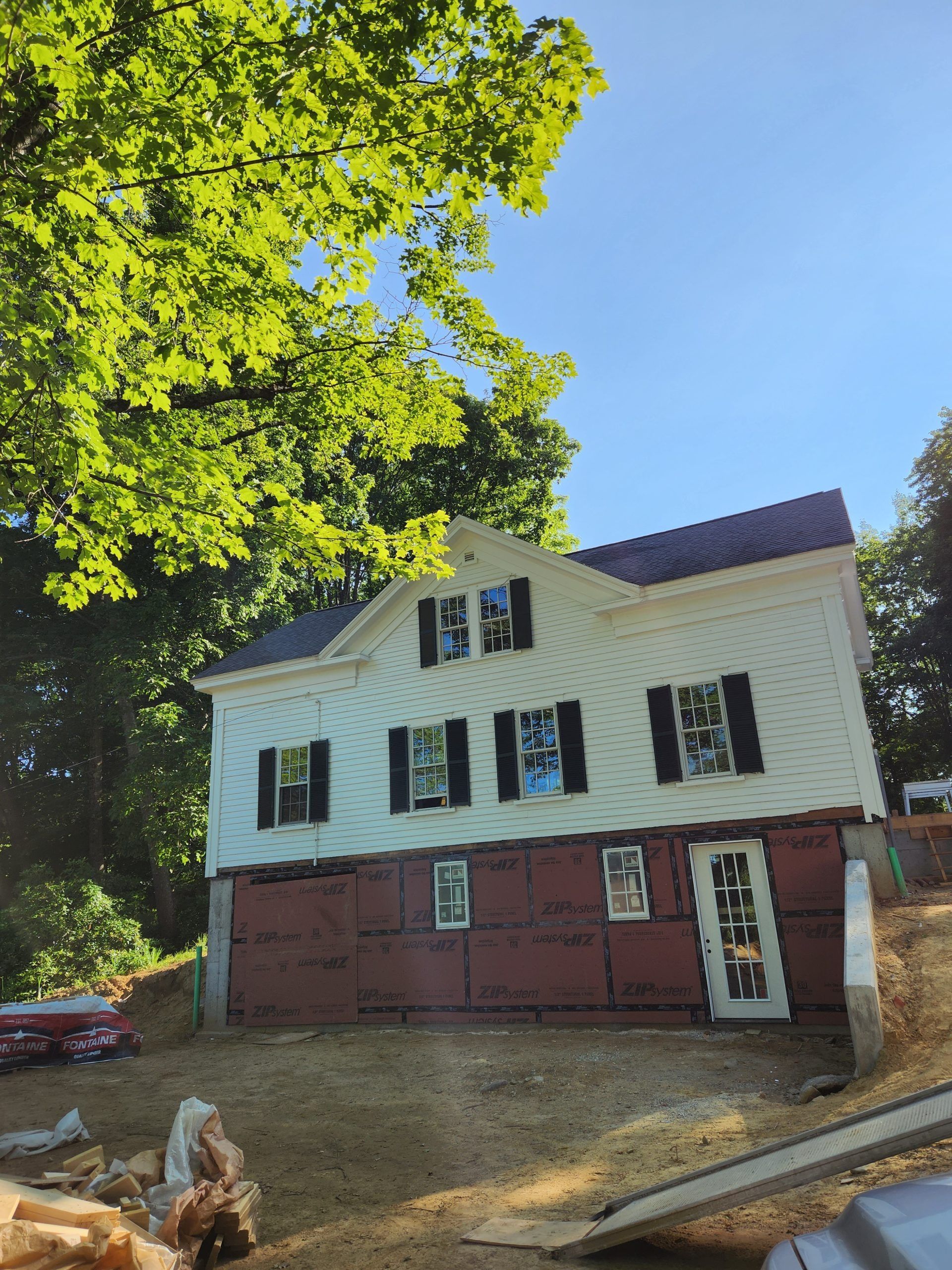 Two-story white house with black shutters, red siding on lower level, under a tree on a sunny day.
