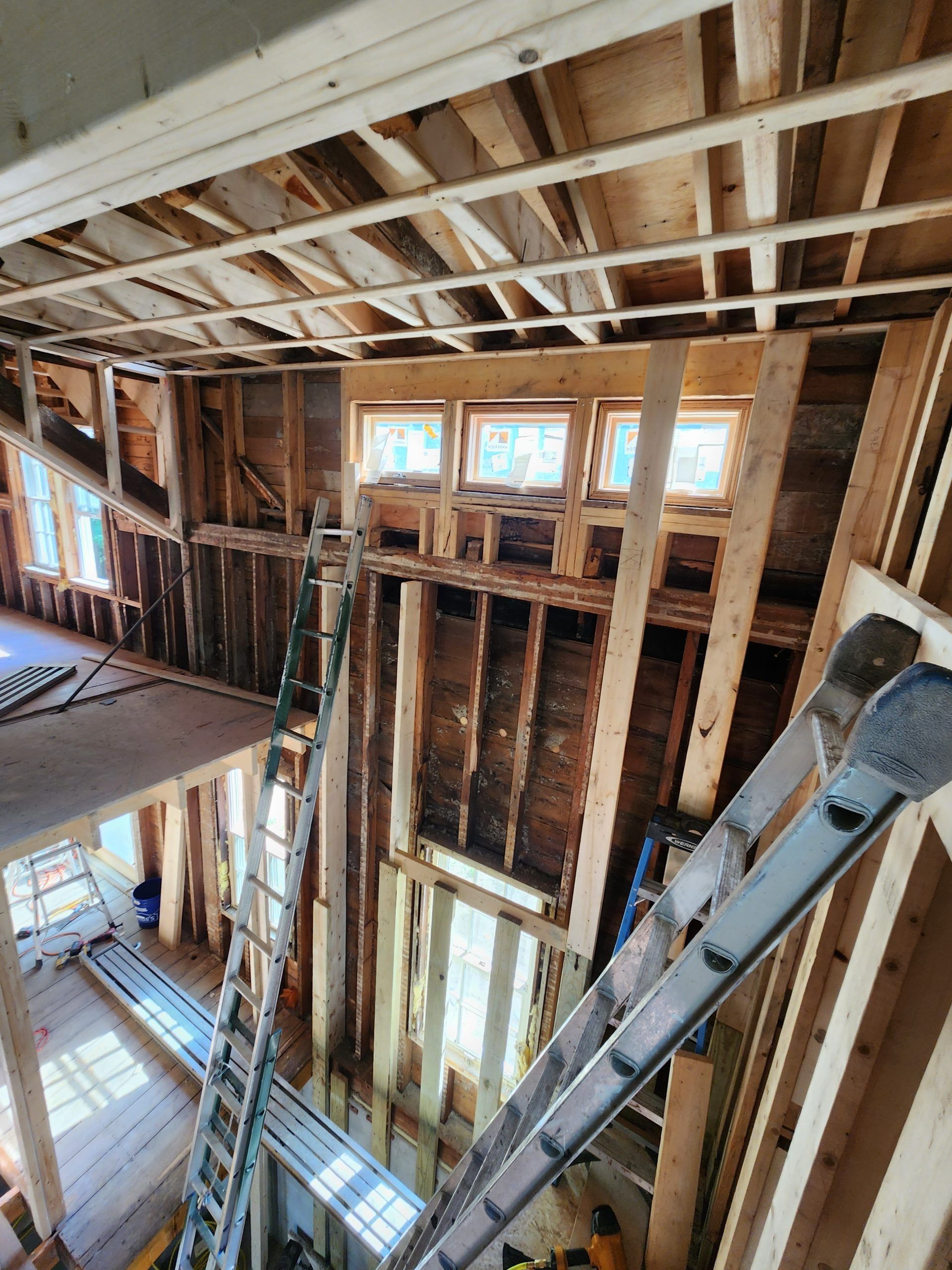 Interior house framing with three windows, two ladders, and exposed wooden structure during renovation.