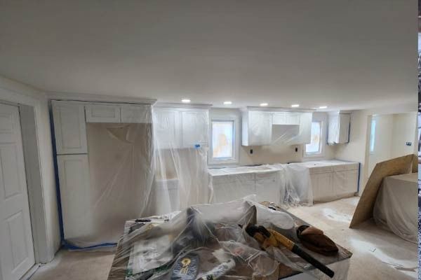 Kitchen under renovation, white cabinets and counters covered with plastic, tools on a work table.