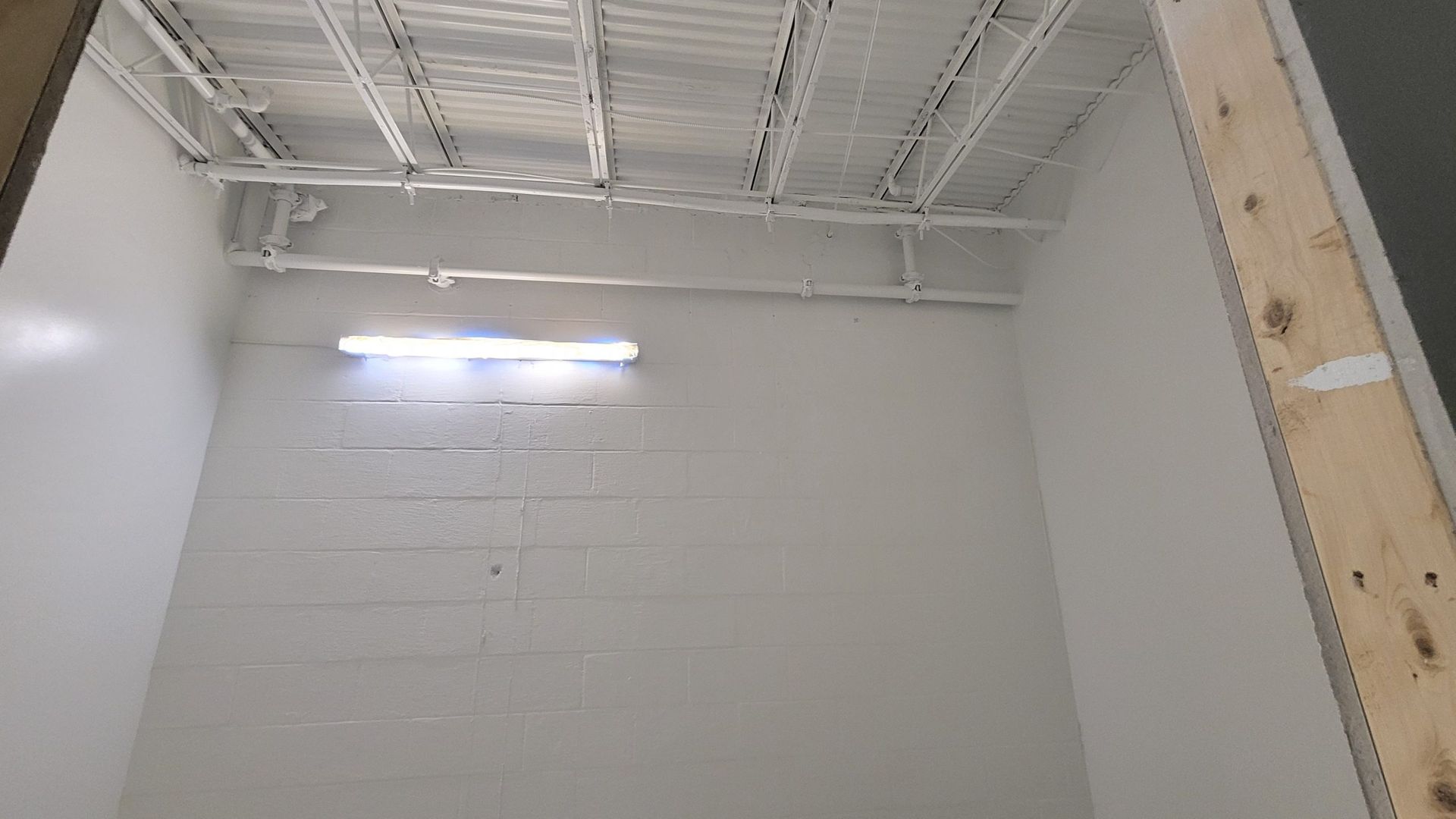 White-painted interior room with exposed ceiling, a horizontal light fixture on the wall, and a wooden frame visible.