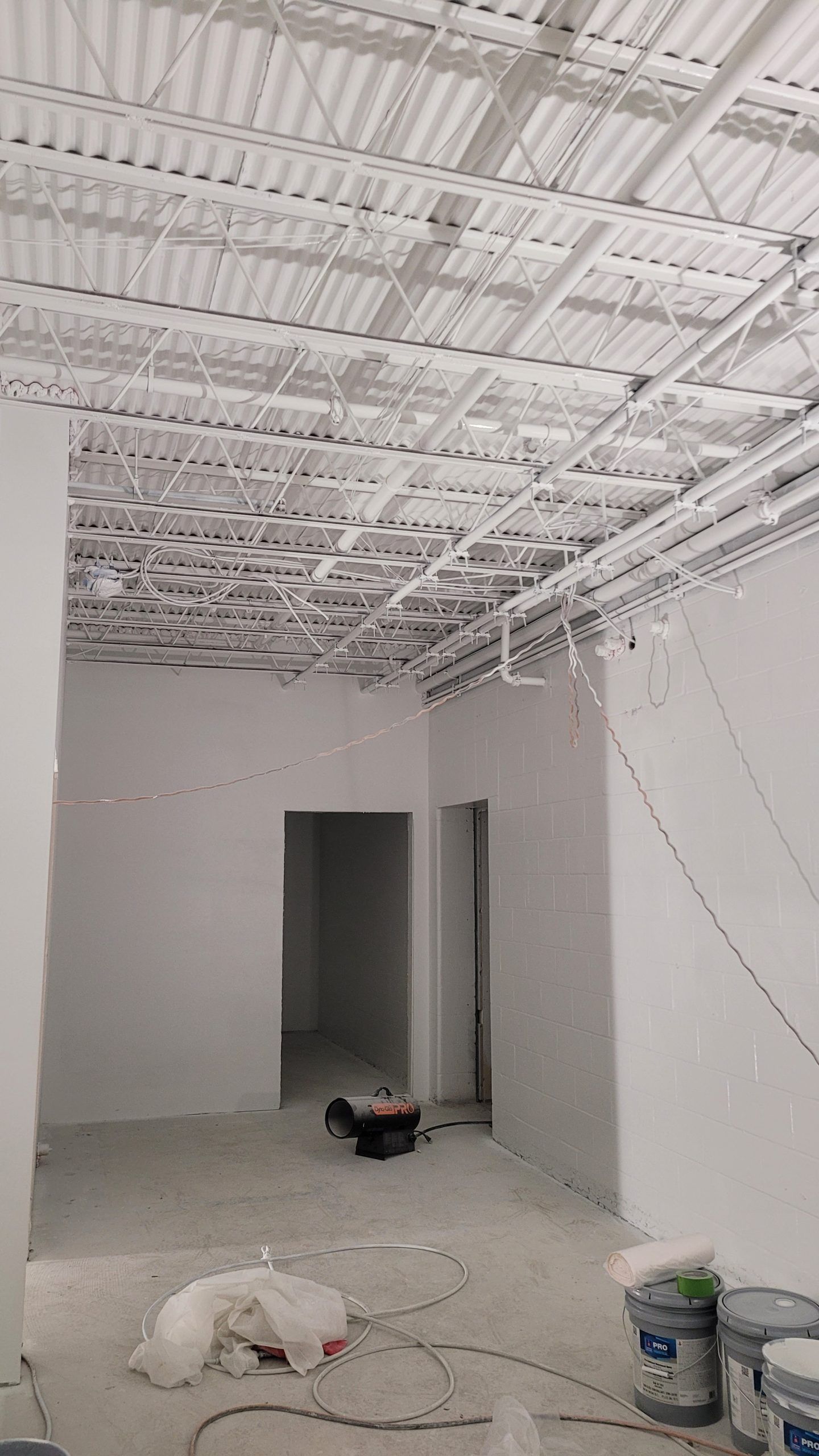 Interior view of a room under construction with white walls, ceiling, and exposed pipes.
