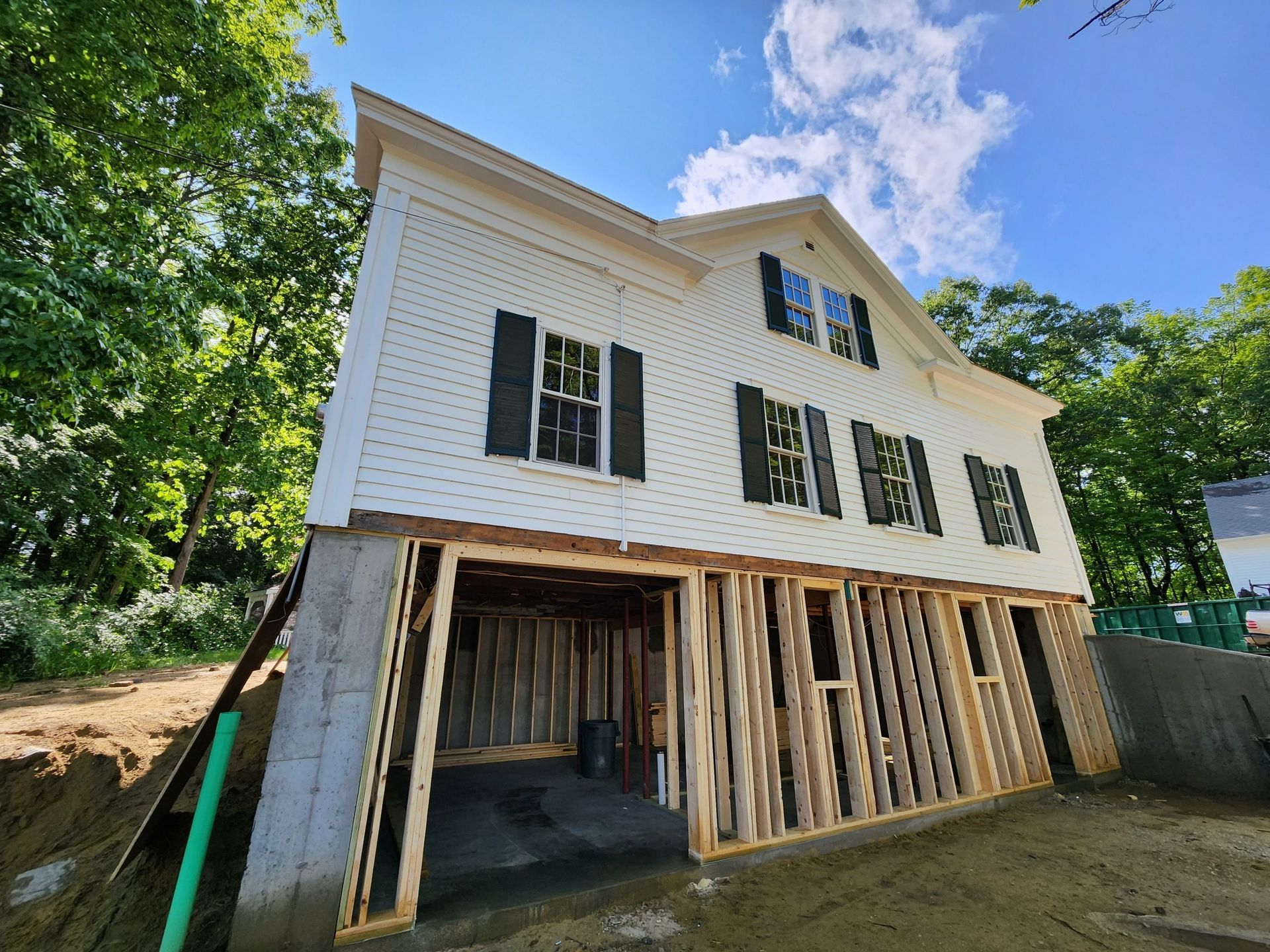 Two-story white house under construction, exposed wood framing at ground level, surrounded by trees and blue sky.