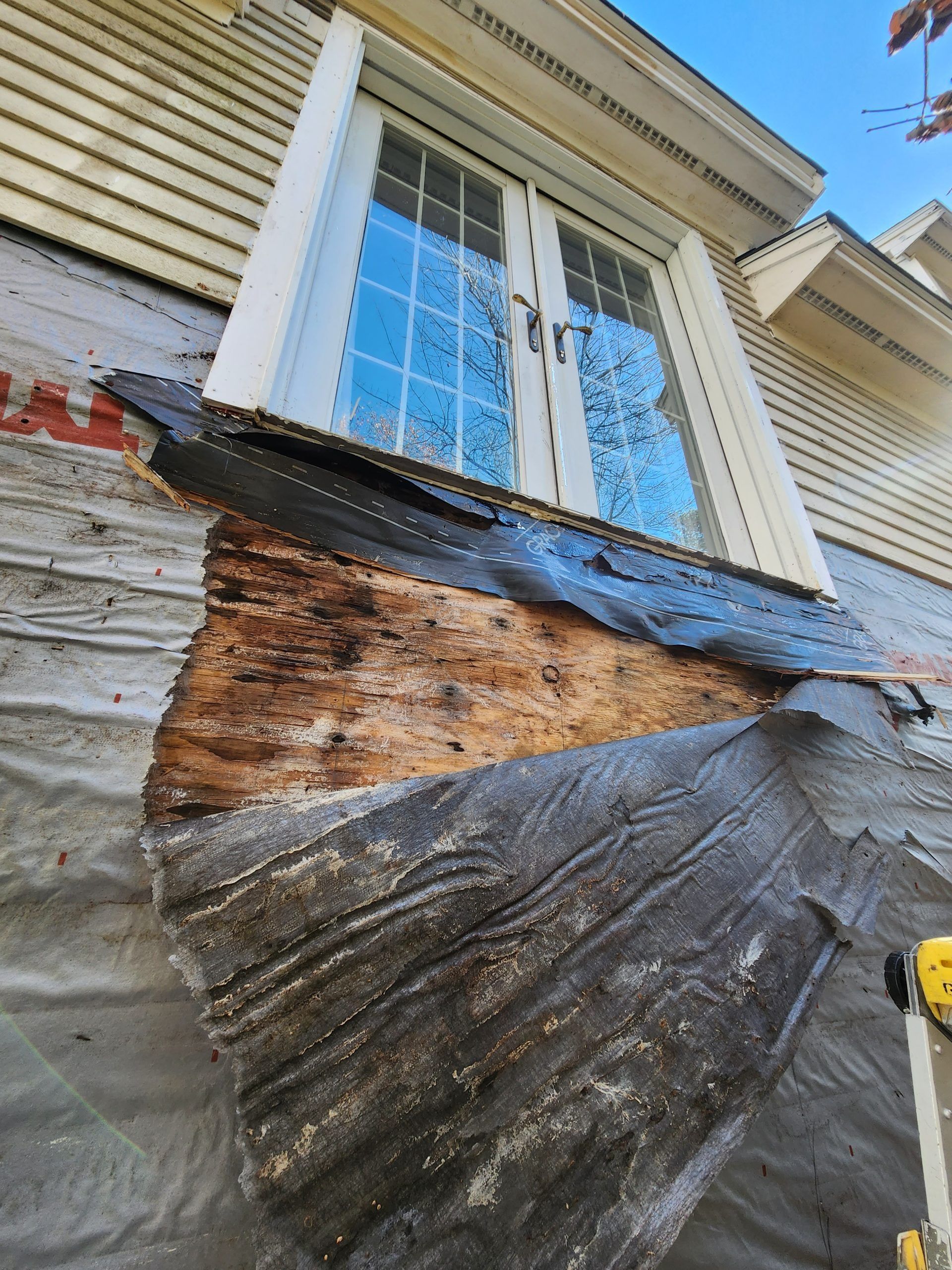 Damaged siding near a window, revealing rotted wood. Black roofing material and gray wrap are visible.