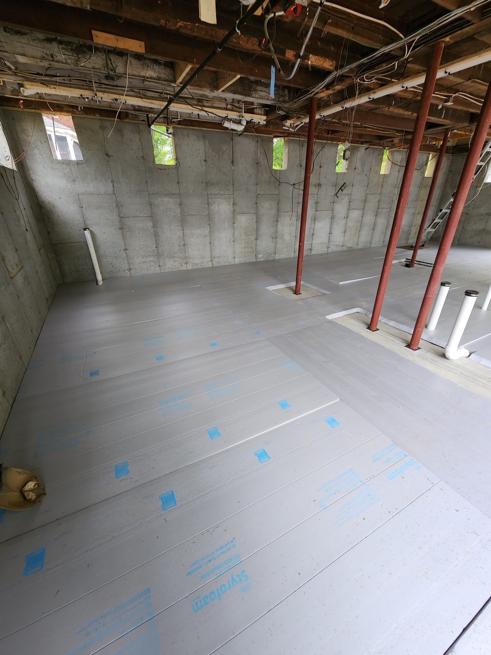 A basement with grey floor membrane, concrete walls, support beams, and plumbing.