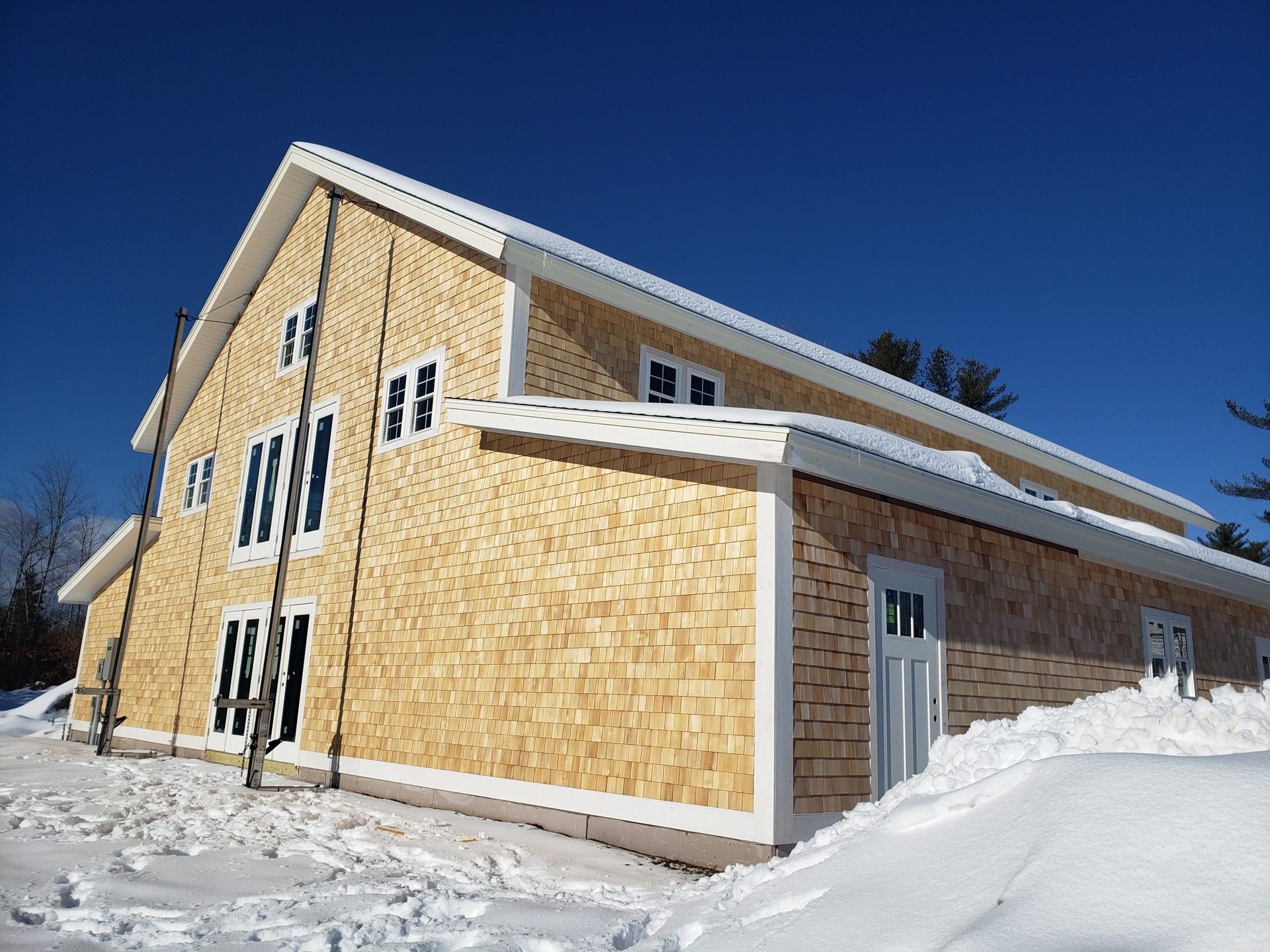Two-story wood-shingled building under construction, white trim, snow on ground and roof, clear blue sky.