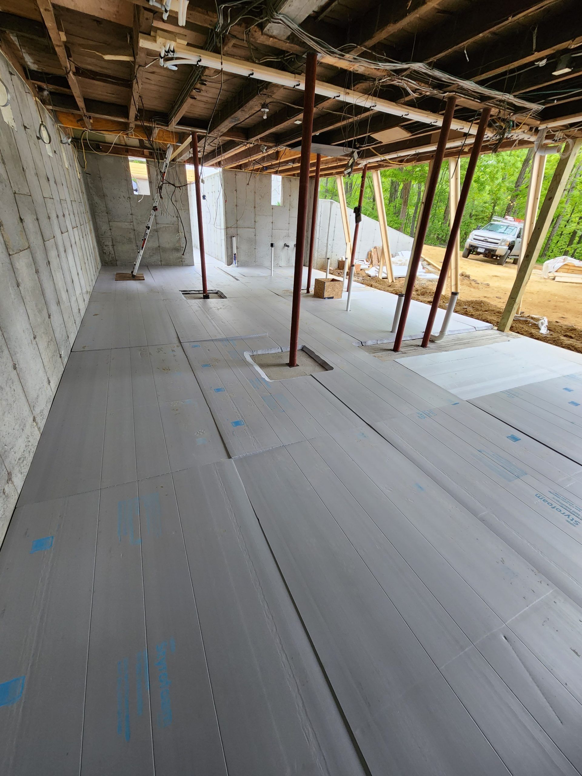 Interior construction site with gray insulation on the floor, support beams, unfinished ceiling, concrete walls, and outdoor view.