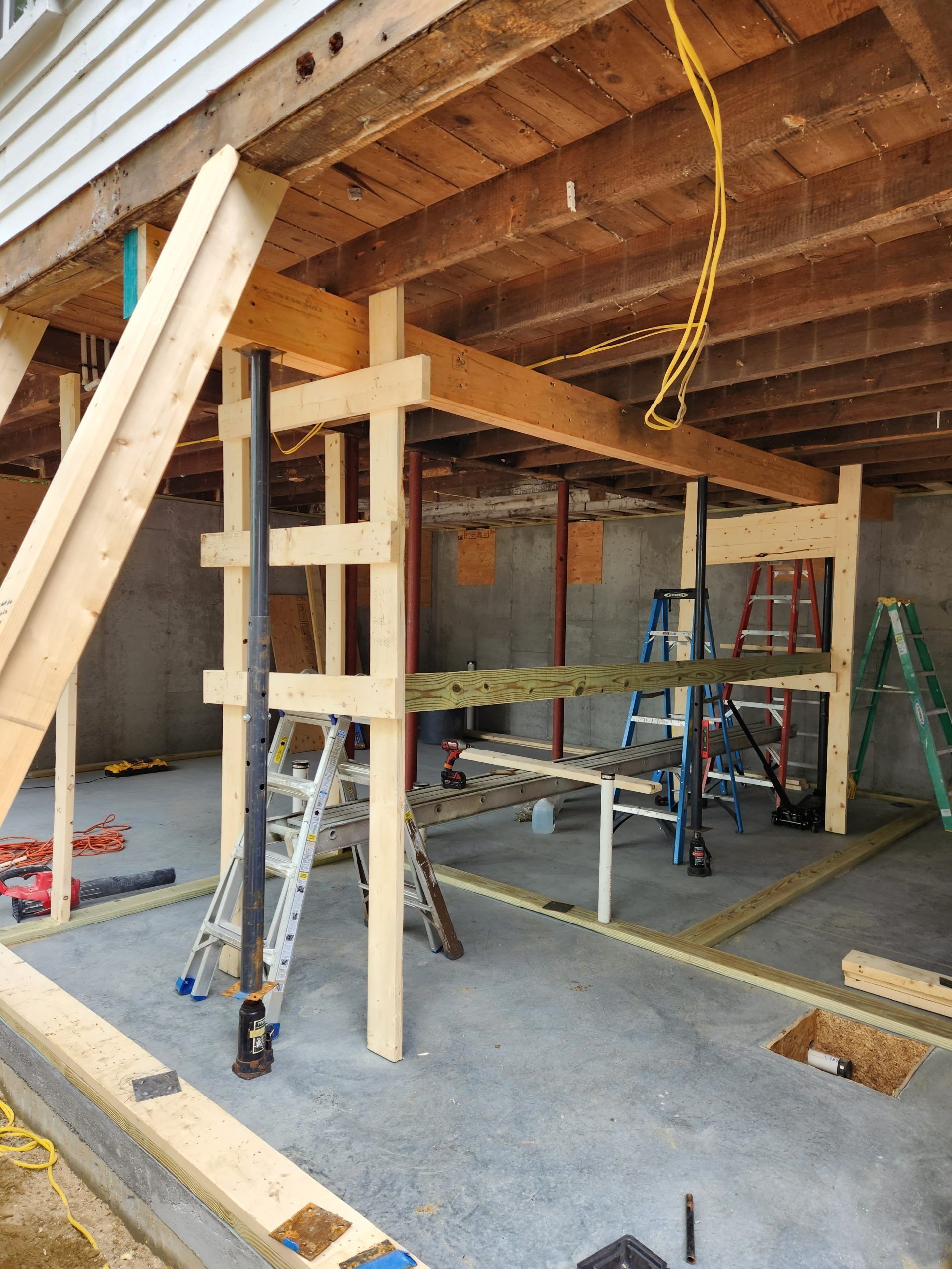 Construction site with wood supports holding up a structure; concrete floor; ladders and tools visible.