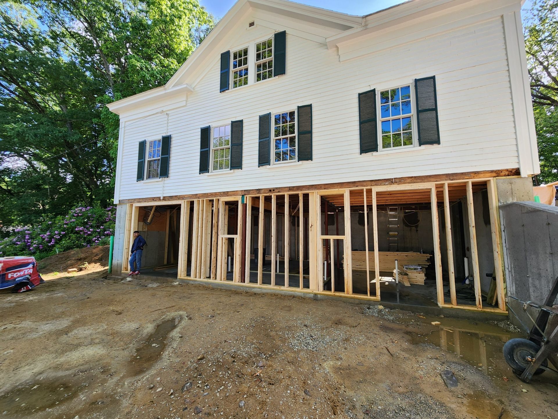White house under construction, exposed wooden framing, dirt yard, green trees in background.