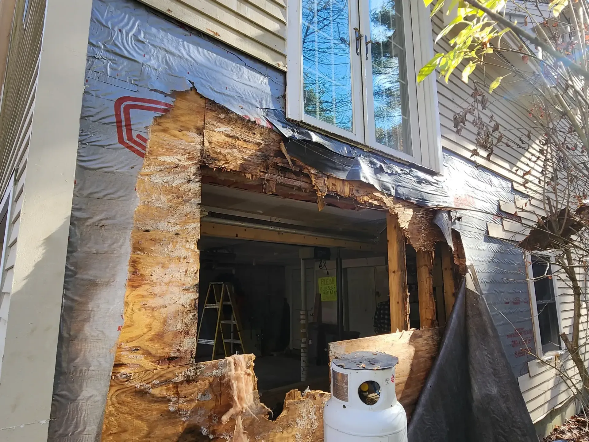 Exterior wall with significant rot around a window. Blue window reflects sky. Propane tank sits below.
