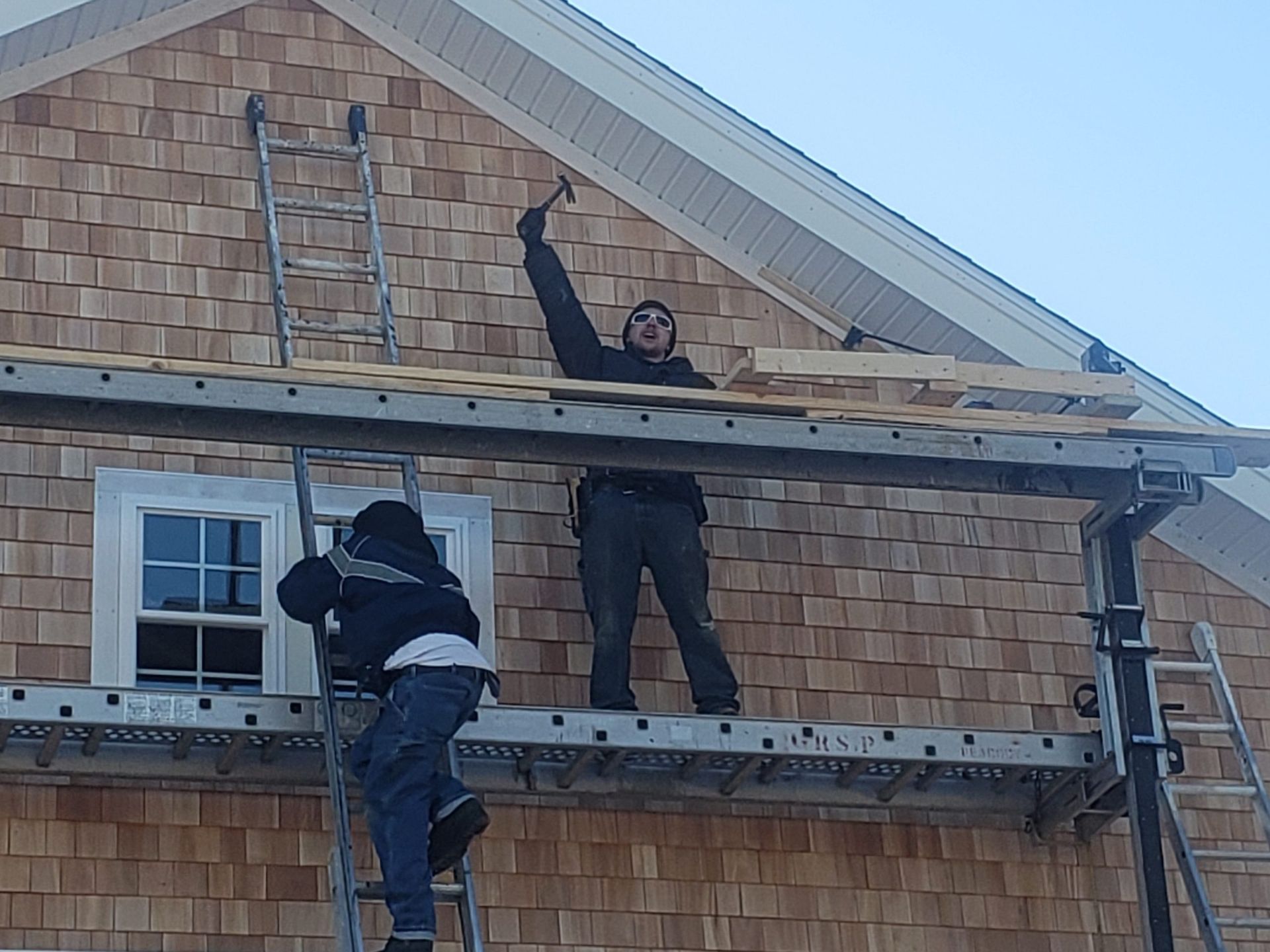 Two people work on a house exterior. One on scaffolding uses a hammer. Another climbs a ladder. Wooden shingles are visible.