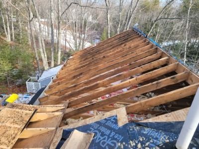 View of a roof in progress, showing exposed rafters, blue underlayment, and surrounding trees.