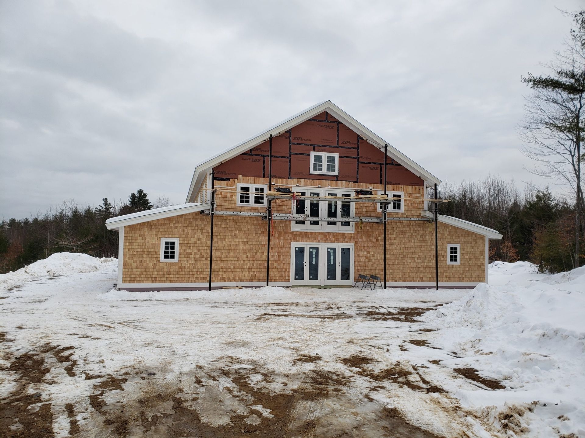 Unfinished two-story house with wood siding, windows, and doors, surrounded by snow and a cloudy sky.