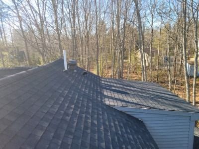 View of a dark shingle roof with a chimney and part of a blue-sided building; trees in the background.