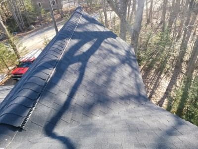 Close-up of a dark asphalt shingle roof, angled, with trees visible, sunny day.