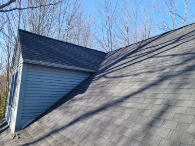 Gray asphalt shingle roof on a house, angled against a blue sky, with shadows and a gray siding wall.