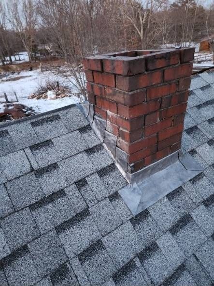 Brick chimney on a gray shingle roof with metal flashing; snow in background.