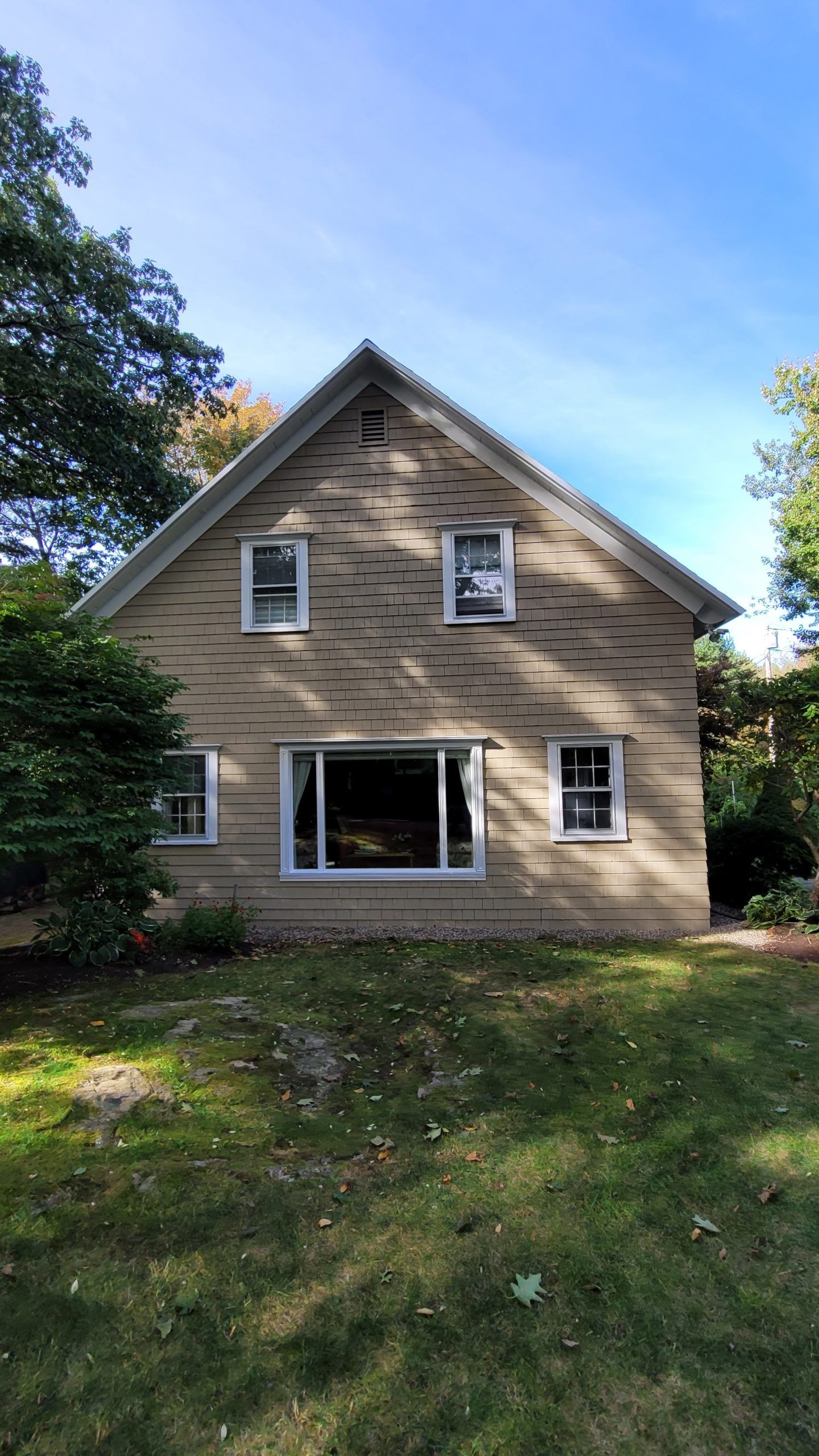 Two-story house with tan facade, white-framed windows, and a peaked roof, set amongst trees on a sunny day.