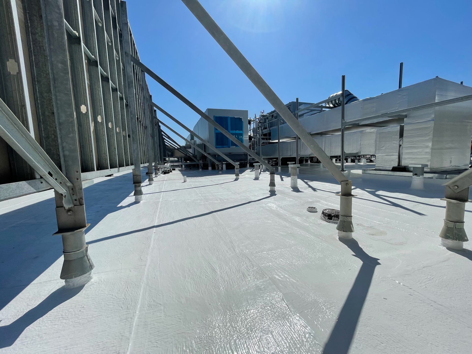 A white commercial roof surface with support scaffolding and a distant HVAC unit under a clear blue sky.