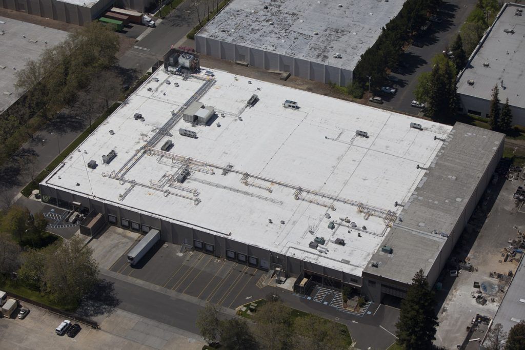 An aerial view of a rectangular industrial building with a white flat roof, surrounded by parking lots and trees.