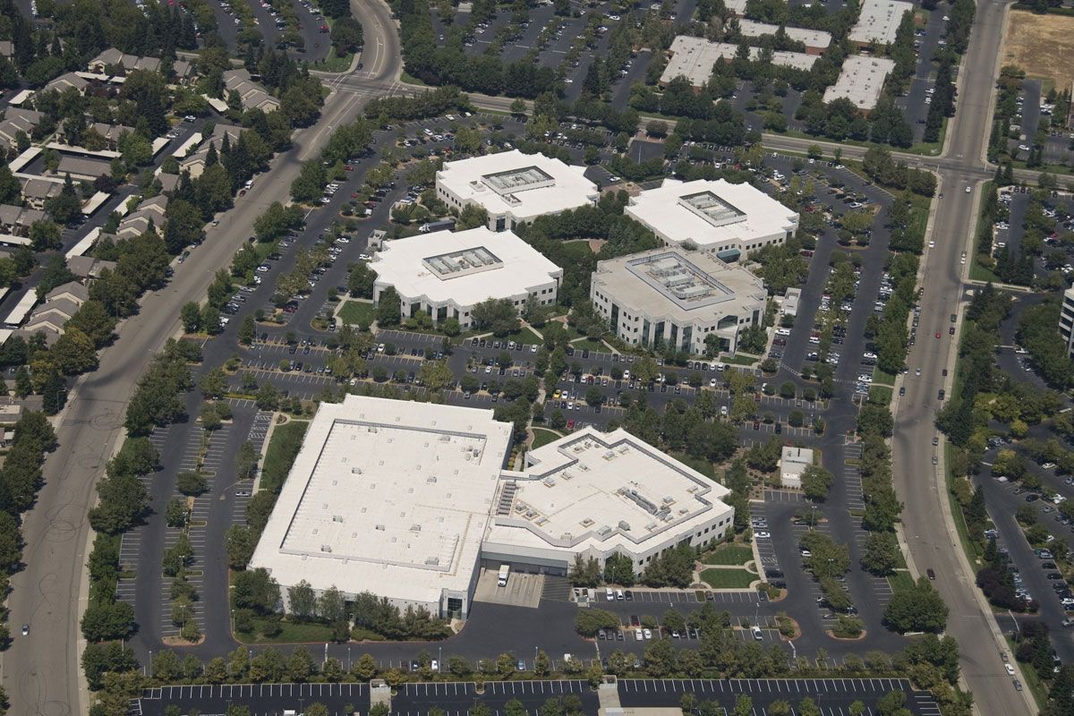 Aerial view of a commercial office park with several interconnected white buildings surrounded by parking lots and trees.