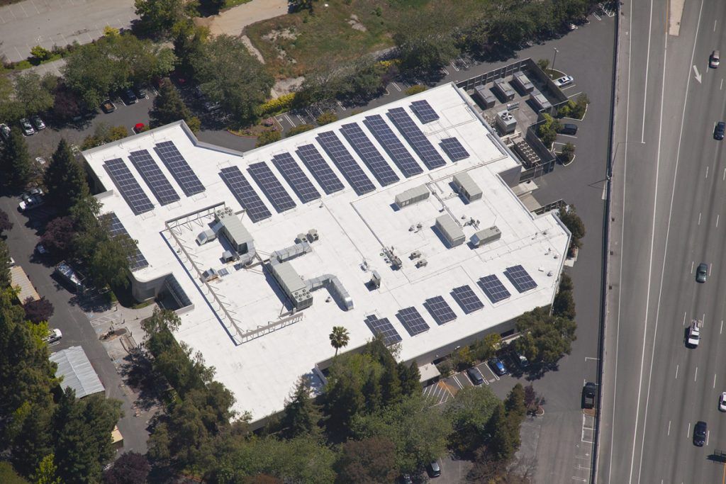 An aerial view of a flat-roofed building featuring several rows of solar panels, surrounded by trees and a parking lot.