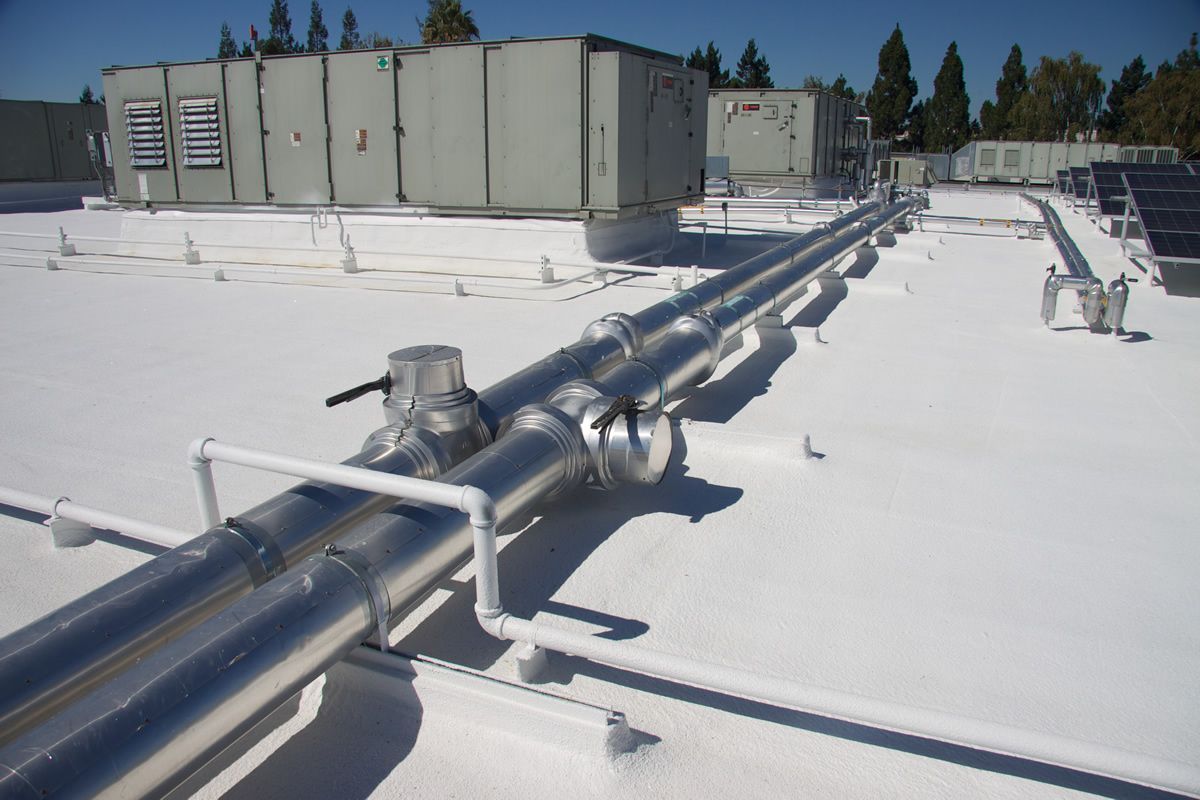 Two insulated metal pipes run across a white rooftop near HVAC units and solar panels.