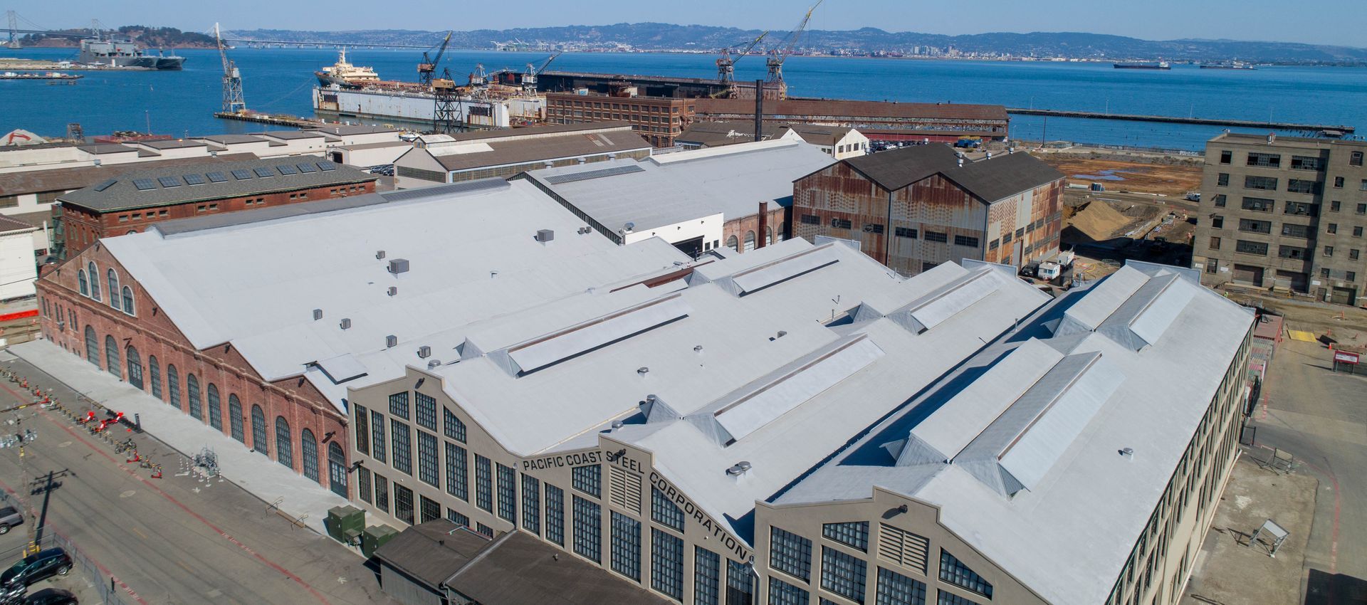 An aerial view of large industrial warehouse buildings with saw-tooth roofs beside a bay with ships and a distant city.