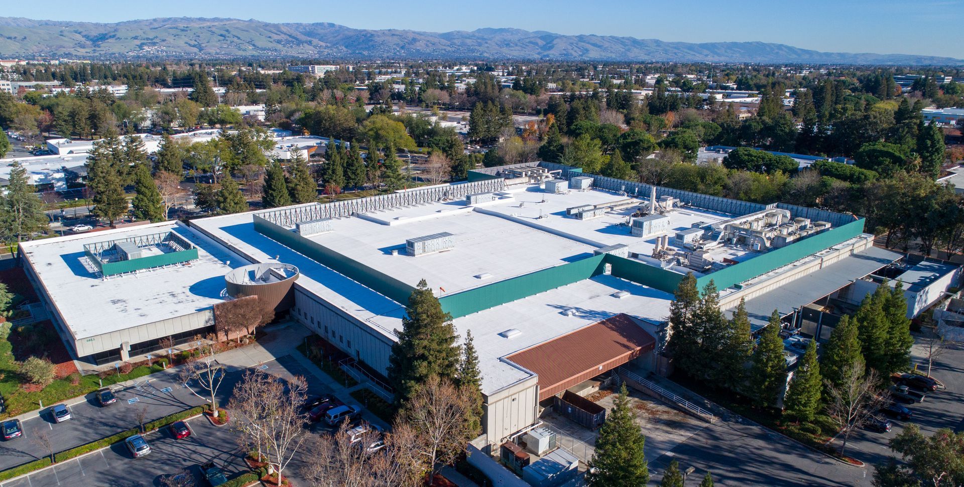 Aerial view of a large flat-roofed commercial building complex surrounded by parking lots and trees under a blue sky.