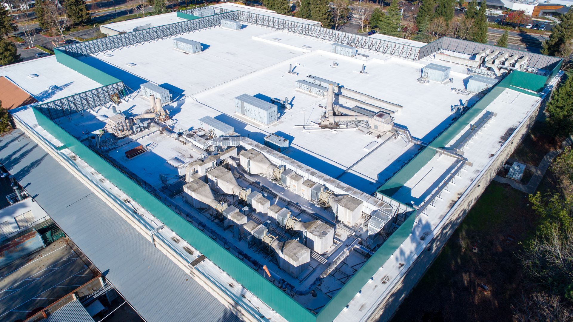 An aerial view of a flat building roof with HVAC units, a green perimeter wall, and a white, reflective surface.