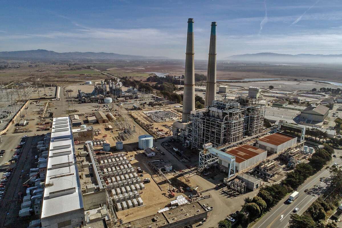 An aerial view of a large power plant with two tall smokestacks, surrounded by industrial structures in an arid landscape.