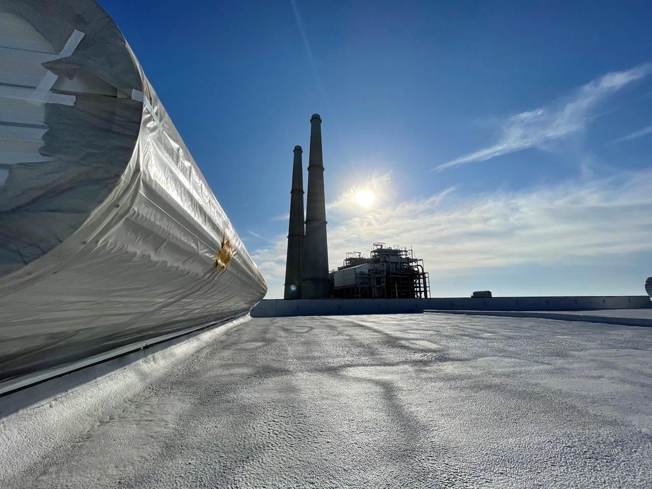 A low-angle view of a flat, white roof leading toward a distant industrial facility with two tall smokestacks under sun.