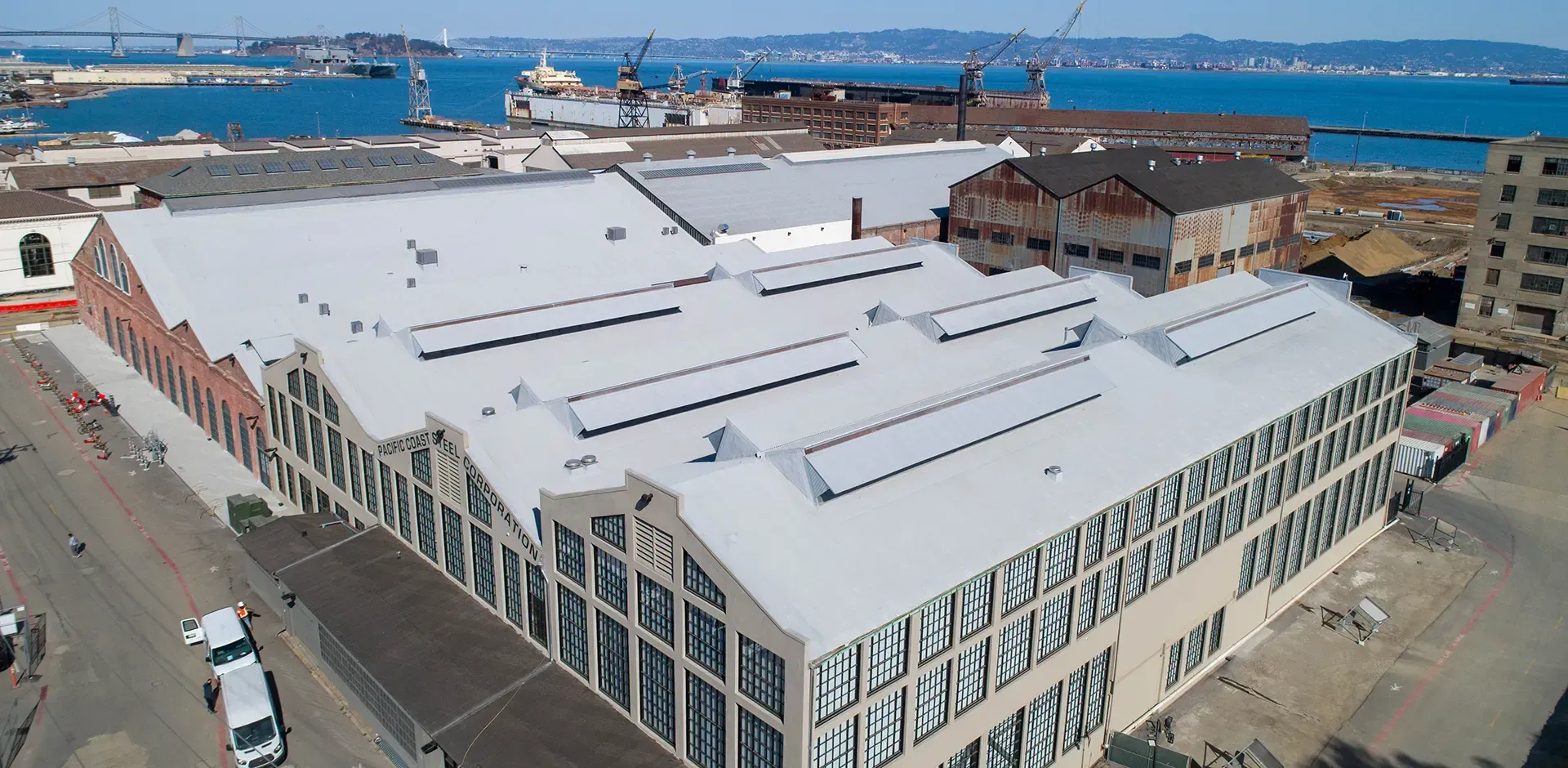 Aerial view of a large industrial warehouse with a light-colored saw-tooth roof, located near a waterfront with cranes.
