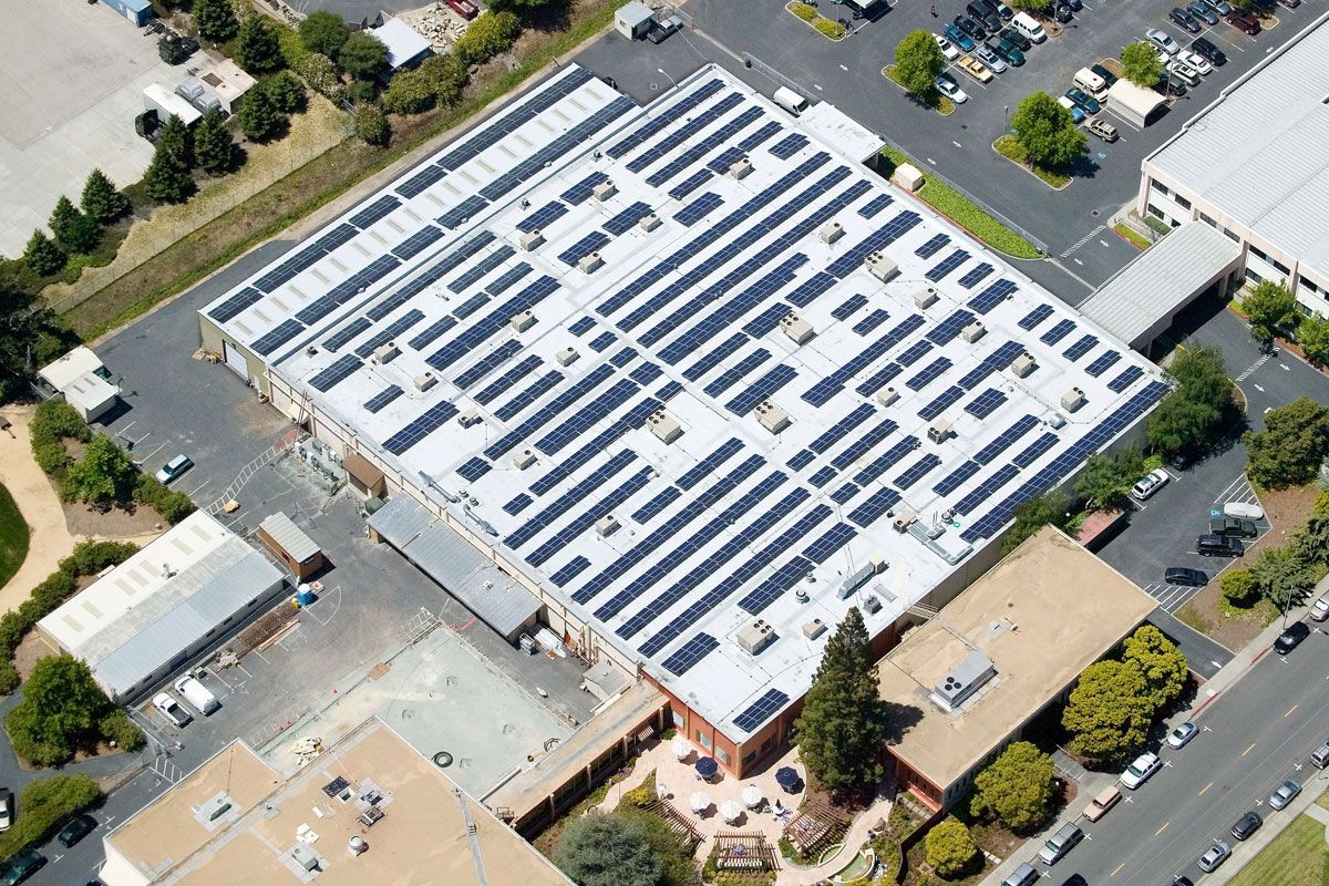 Aerial view of a building rooftop covered in solar panels, surrounded by parking lots and trees.