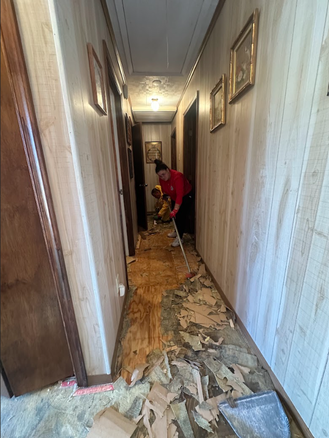 A person in a red shirt uses a broom to sweep debris off a wooden subfloor in a hallway with wood-paneled walls.