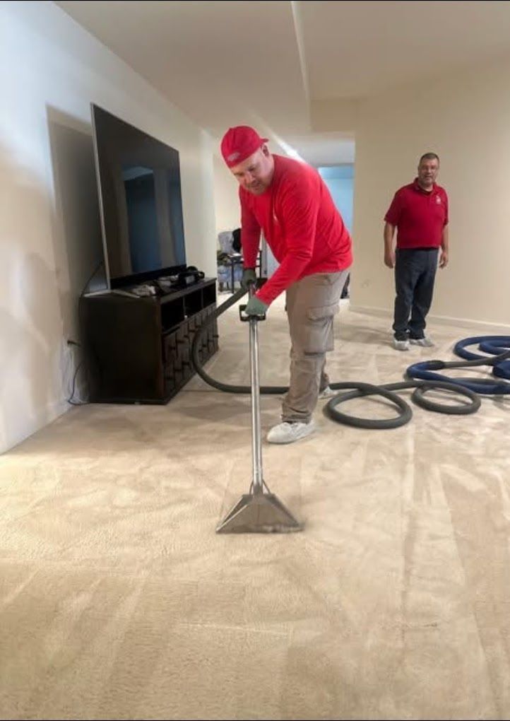 Two workers in red shirts clean light-colored carpet in a living room with a television.
