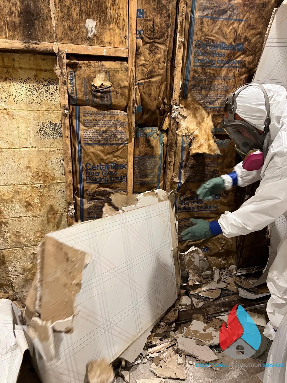 A person in a protective suit and mask removes damaged drywall and insulation from a wall in a water-damaged room.