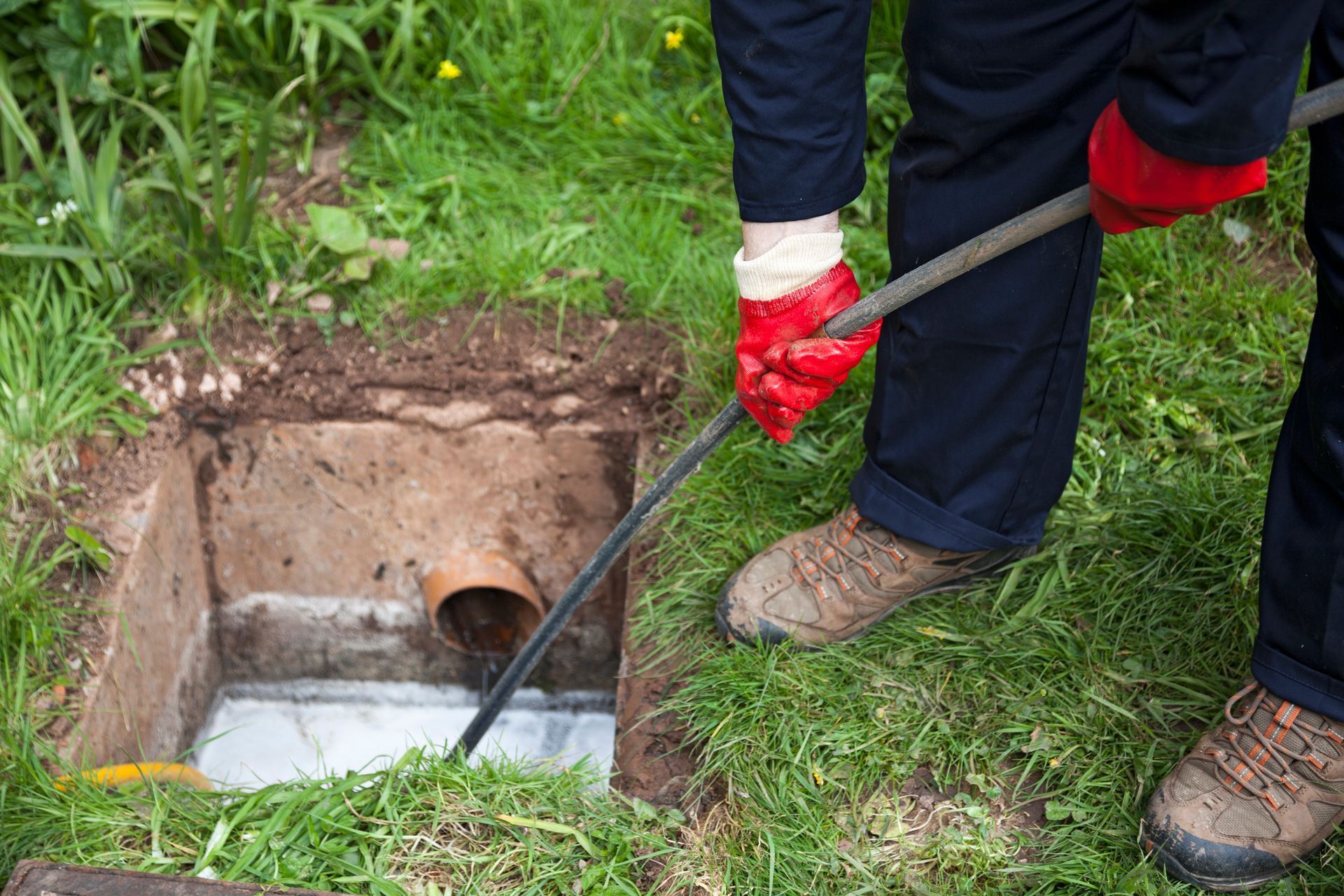 A worker in gloves uses a drain snake to clear a clog in an outdoor sewer access pit.