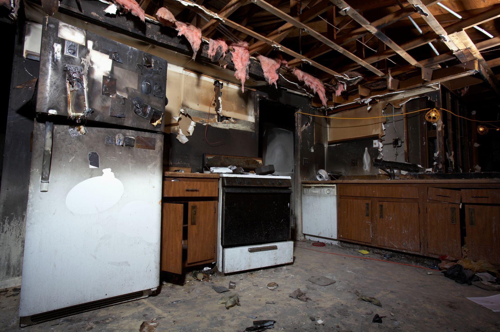 A fire-damaged kitchen interior showing a charred refrigerator, stove, cabinets, and exposed ceiling joists with insulation.