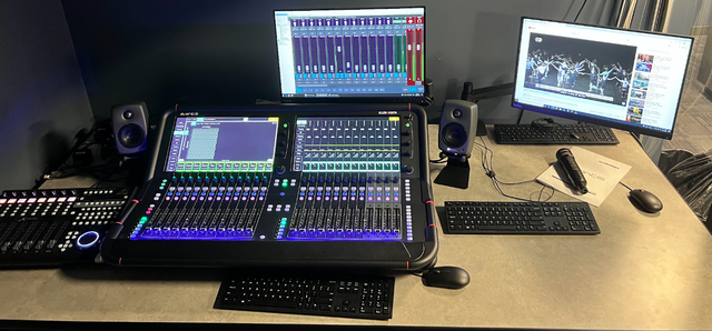 A desk audio mixer with Dante network interface card and a computer , keyboard , monitors and speakers on it.