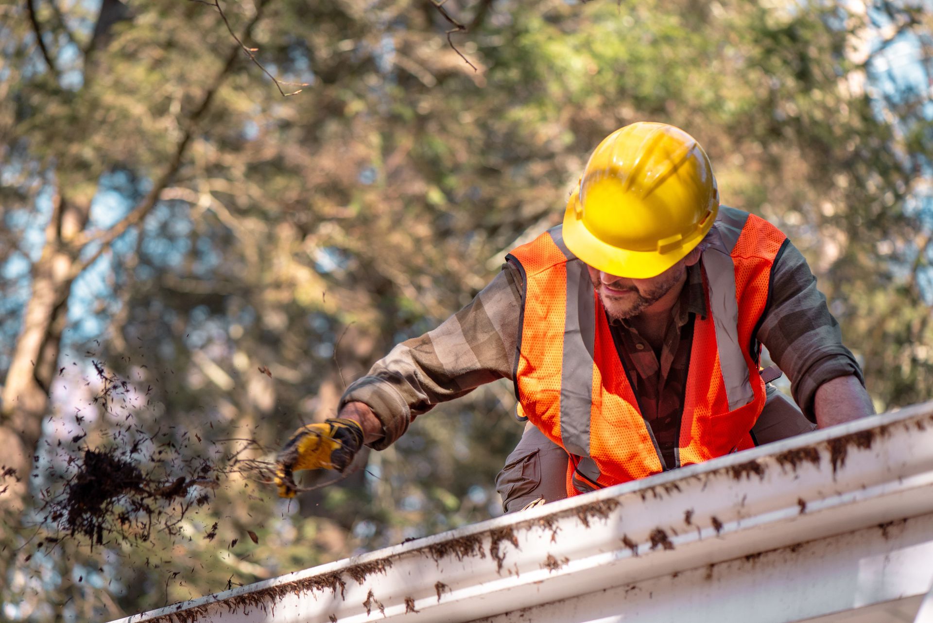 A male contractor wearing a safety helmet and high-visibility vest cleans debris from house gutters