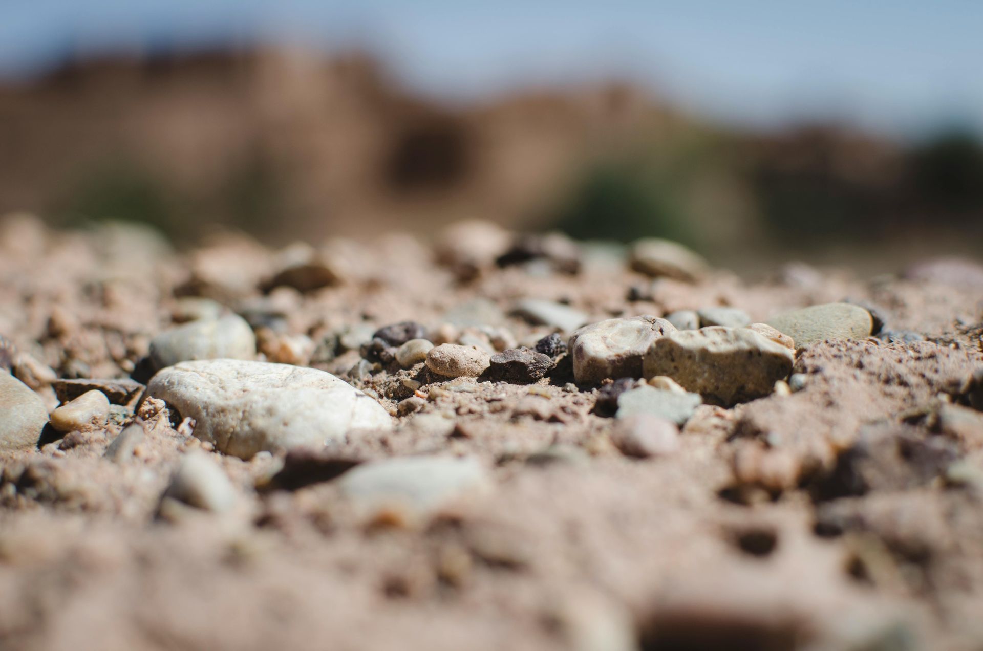 Close-up of a rocky, sandy ground with small stones scattered on the surface. Blurry background of a brown structure.