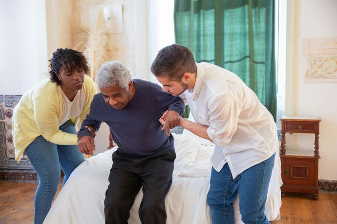 Two people helping an older person sit on a bed; a bedroom with white sheets, curtains.