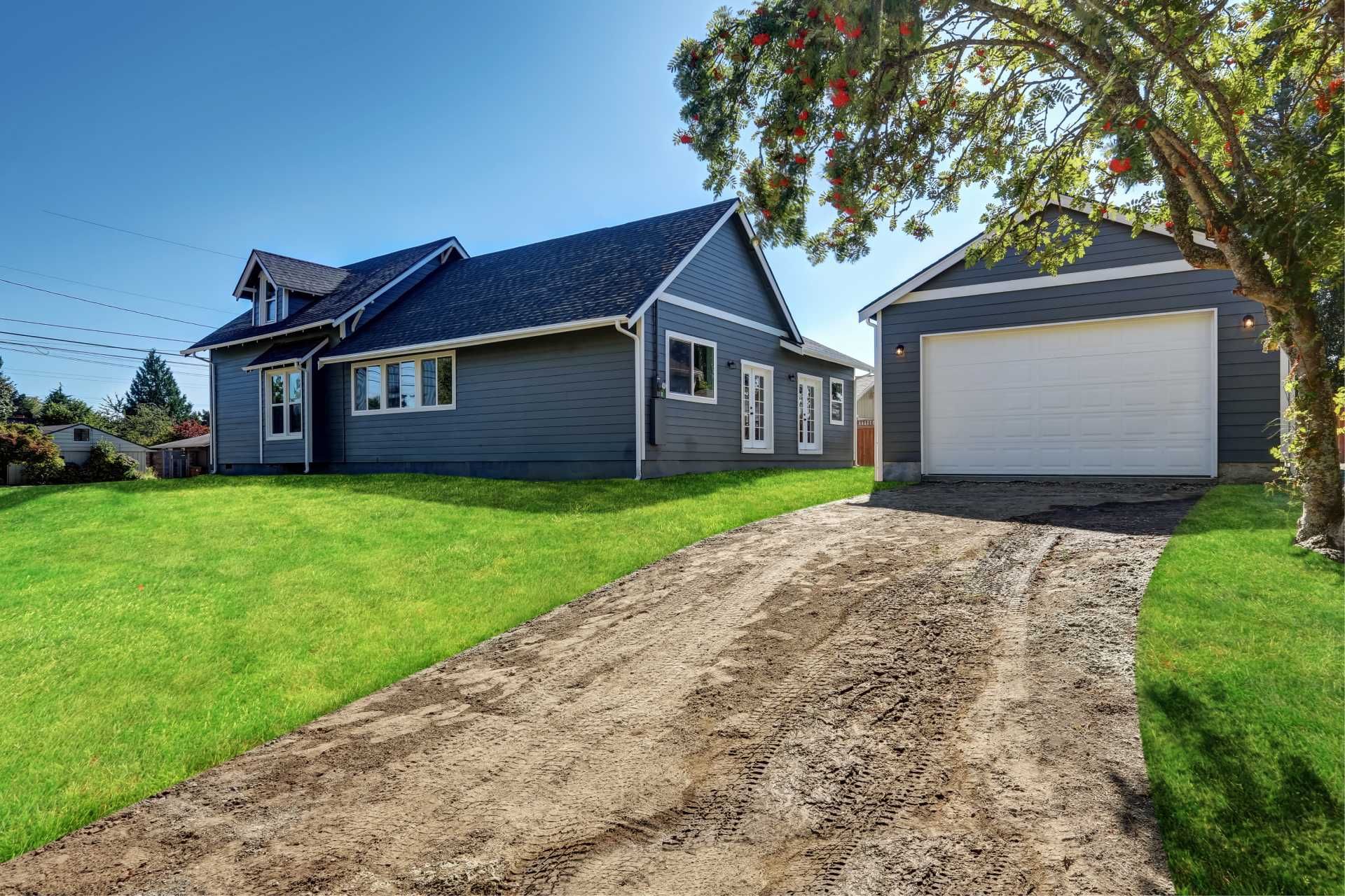 Gray house with attached garage and muddy driveway, green lawn, and blue sky.