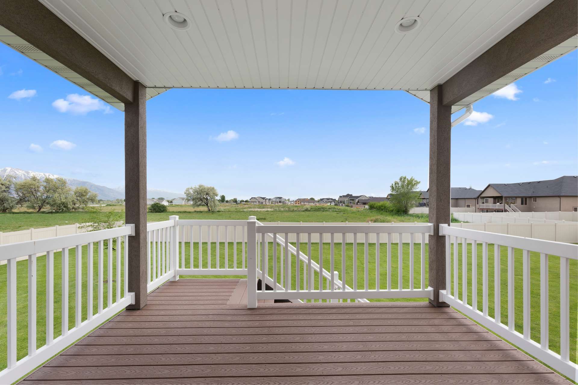 Covered porch overlooking a grassy yard, blue sky, and suburban homes. Brown deck, white railings, and brown beams.