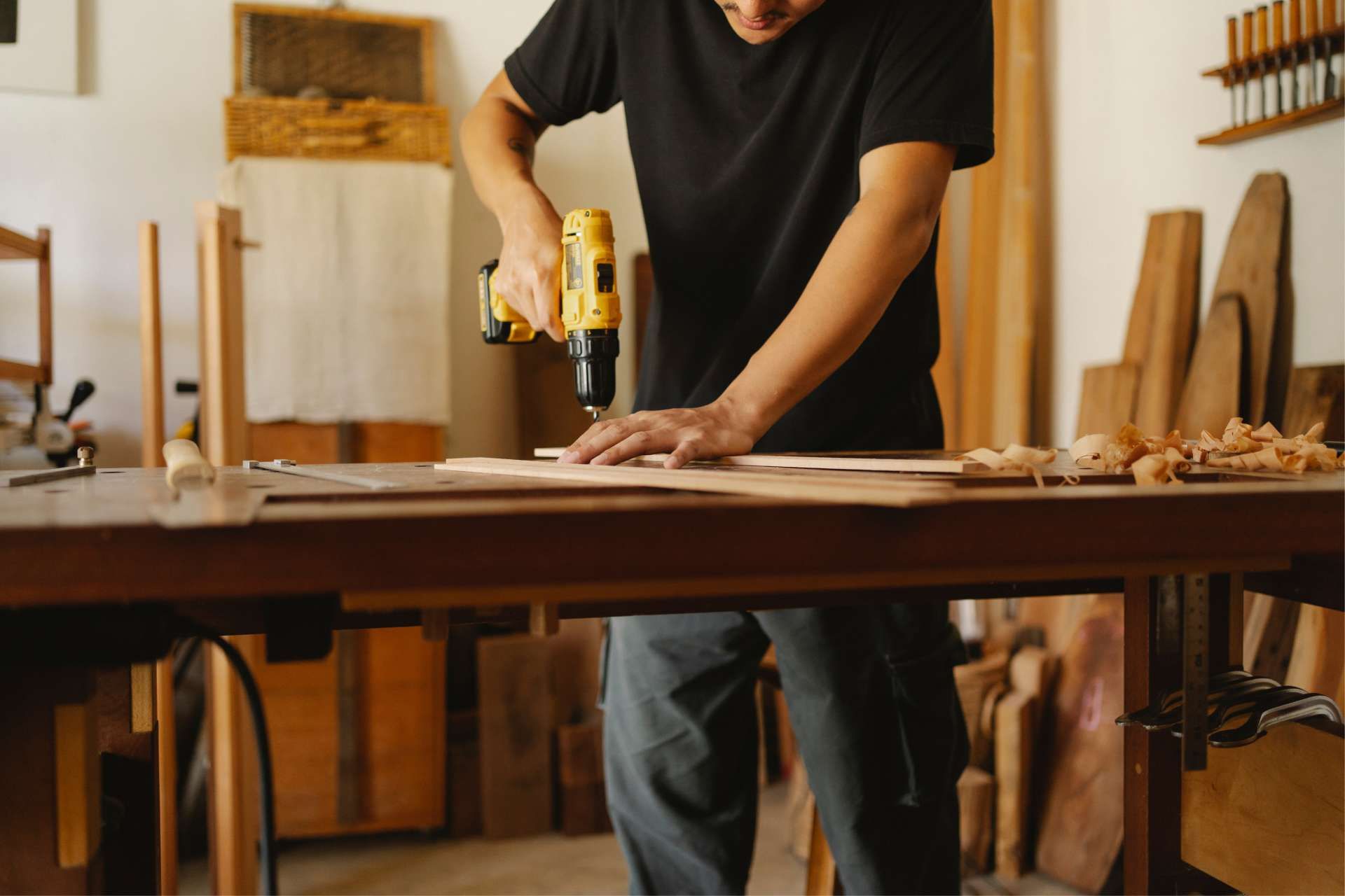 Carpenter using a drill on a wooden plank in a workshop.