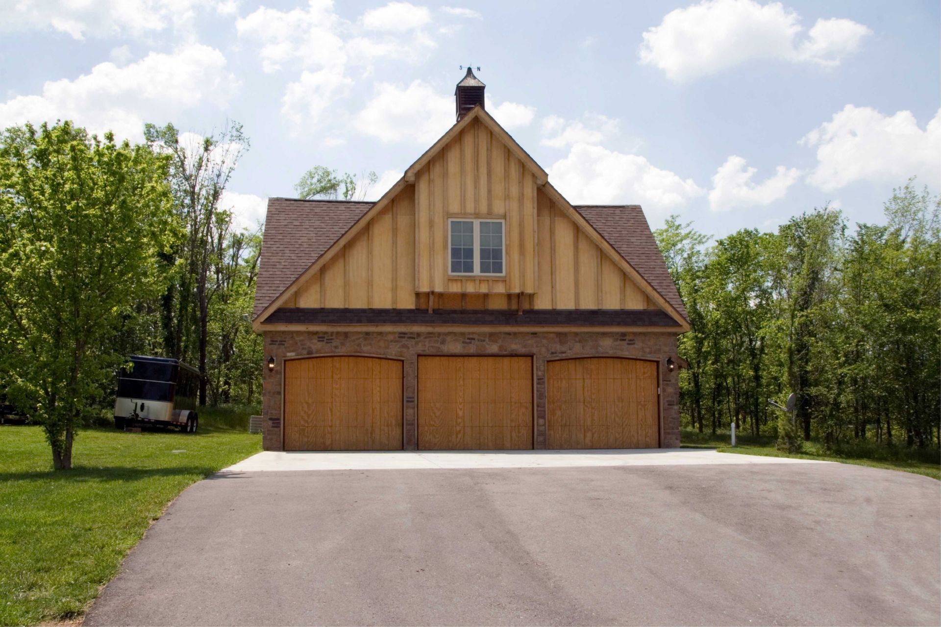 Three-car garage with a brown roof and wooden doors, set on a paved driveway with trees.